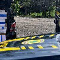 A imagem em destaque mostra uma viatura da Polícia Rodoviária Federal próxima à carga de maços de cigarros contrabandeados apreendida.